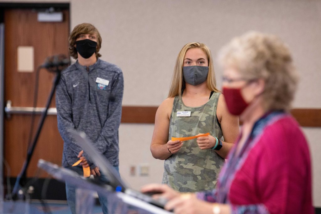 Students is masks standing next to woman speaking into microphone. 