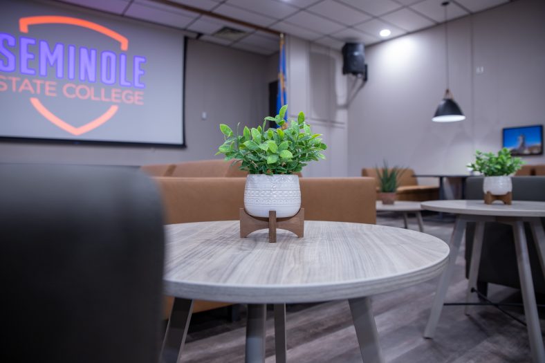 Pictured are several chairs, couches and tables featuring green plants. This seating is in front of a large projector screen above the stage in the new Virtual Learning Lounge.