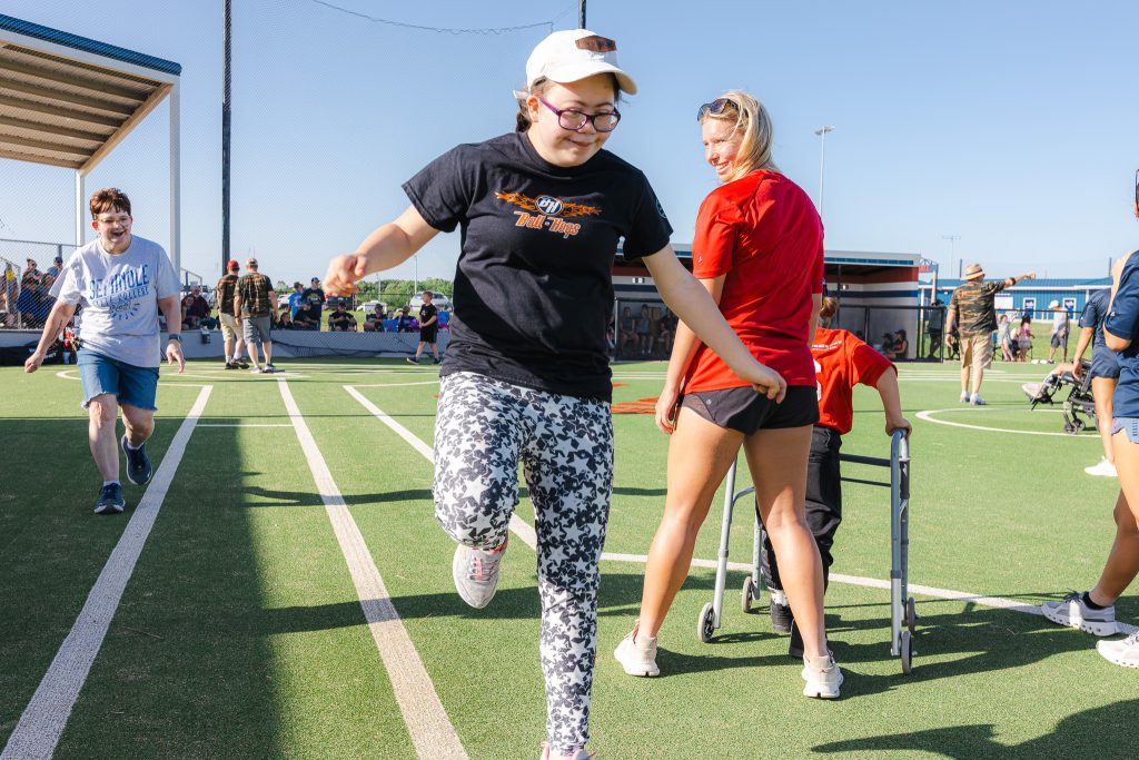 In this photo, A League of Their Own player Hannah Choate skips to first base on Monday, April 27, at the Avedis Foundation Adaptive Field inside the Brian Crawford Memorial Sports Complex. She is cheered on by SSC Student Support Services Advisor Sarah Ledford, left, and Trojan soccer player Bella Mattingly, right.